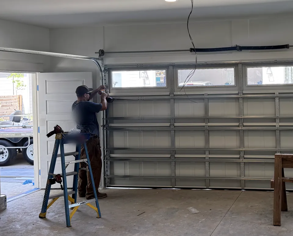A technician on a step ladder fastening the track of a newly hung garage door with a row of windows, inside a newly built garage